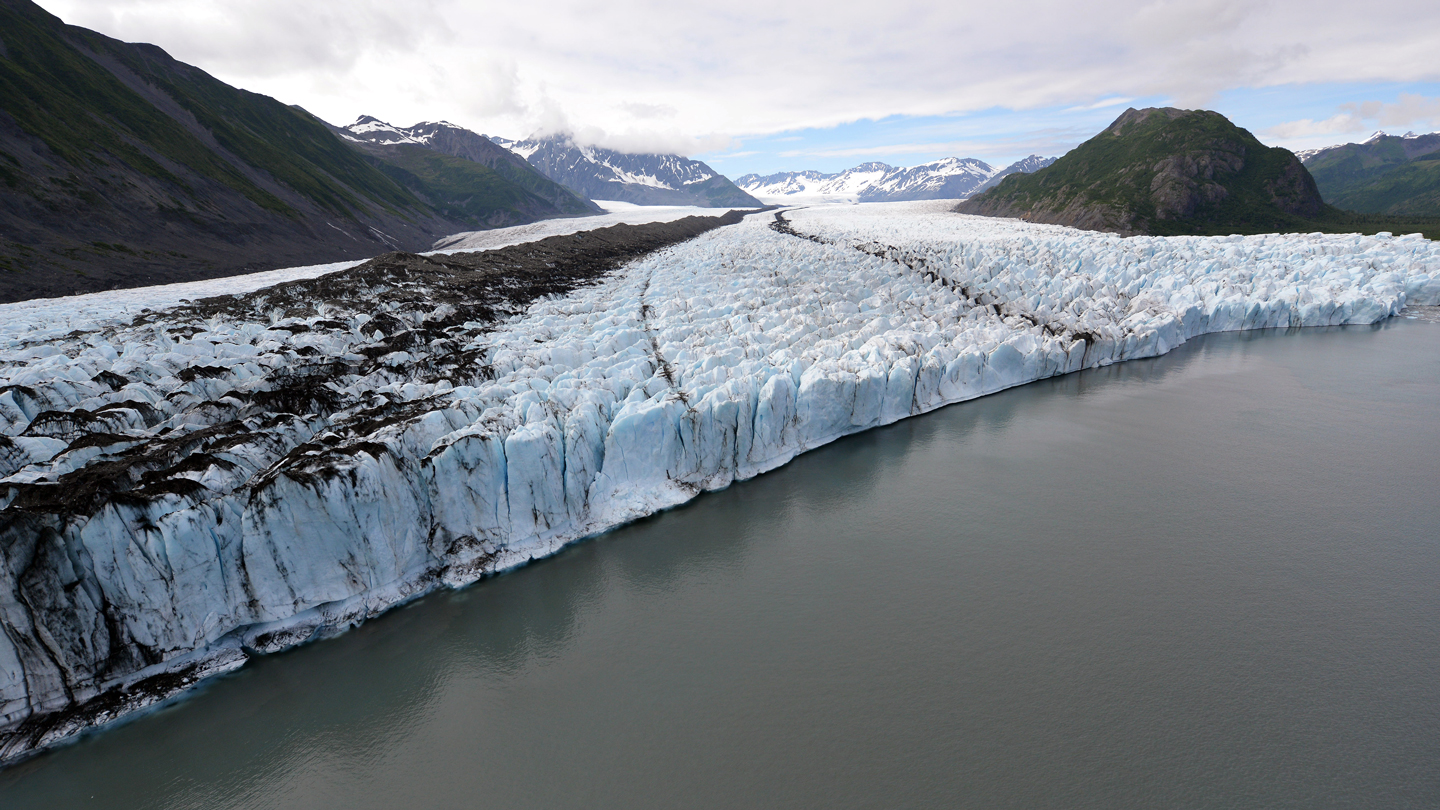 An aerial view shows a wide glacier flowing between mountains and ending at a gray glacial lake.