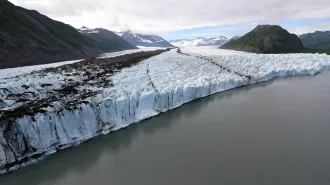An aerial view shows a wide glacier flowing between mountains and ending at a gray glacial lake.
