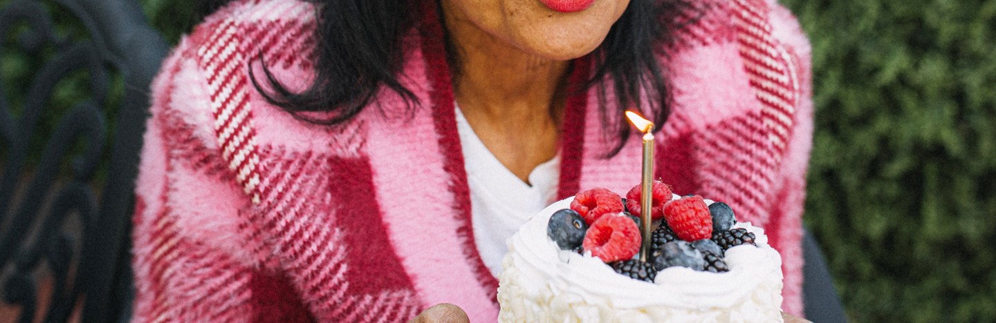 A middle-aged woman in a pink plaid sweater blows on a single, lit candle in a cake covered with white frosting and mixed berries.