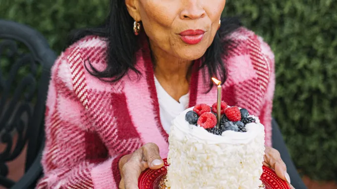 A middle-aged woman in a pink plaid sweater blows on a single, lit candle in a cake covered with white frosting and mixed berries.