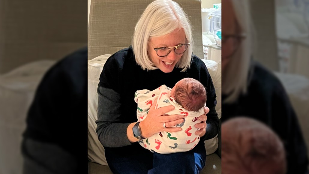 A woman holds a swaddled baby in her lap and looks like she is talking to the infant.