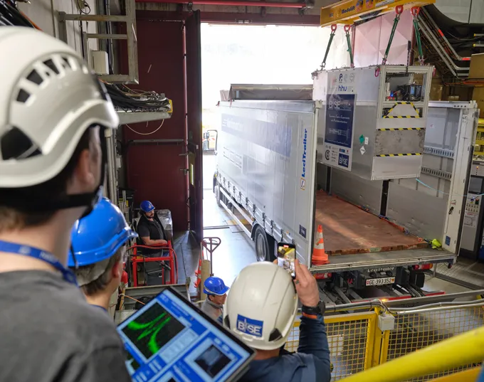 Scientific equipment is lowered by crane into the back of a truck, while people in hard hats look on.