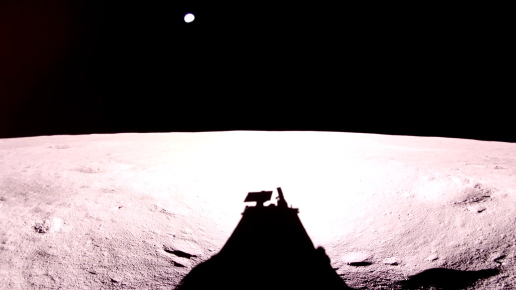A long spacecraft shadow falls across the moon’s surface beneath a black sky.
