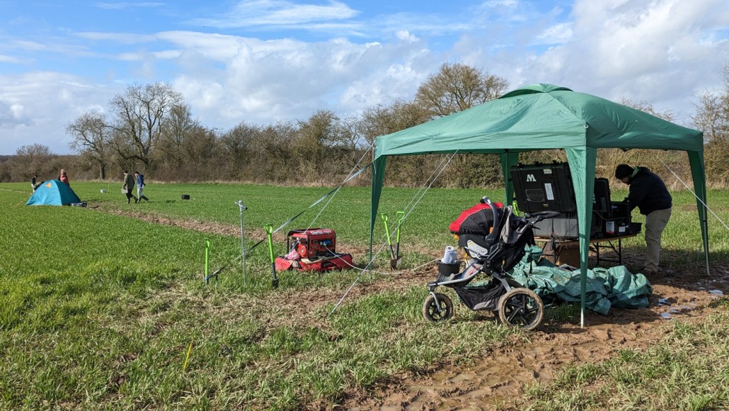 A group of scientists work inside a tent. Nearby, another group is walking in an open, grassy field.