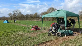 A group of scientists work inside a tent. Nearby, another group is walking in an open, grassy field.