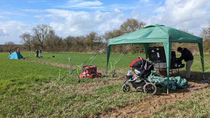 A group of scientists work inside a tent. Nearby, another group is walking in an open, grassy field.