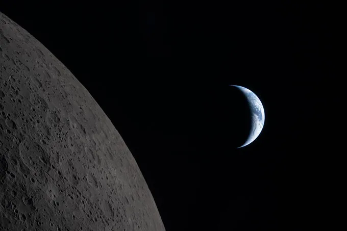 The cratered curve of the moon is in the foreground wiht a blueish crescent of Earth in the background.