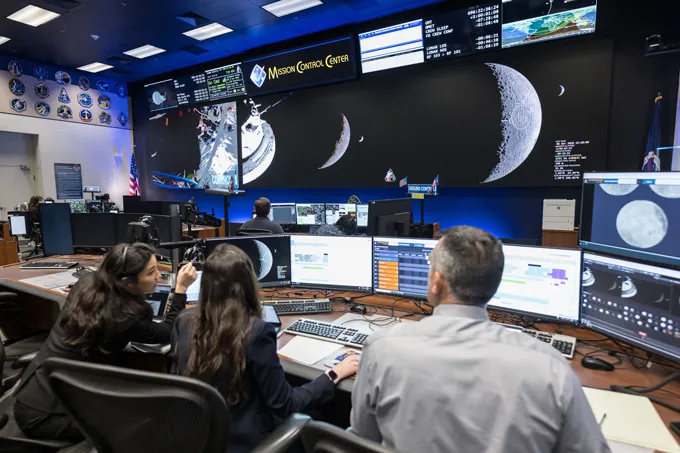 Two women and one man sit at a desk looking at large screens with photos of the moon and a sign reading "Mission Control Center"
