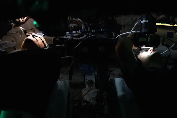 Two astronauts look up in a dark spacecraft. The only light is on their faces. Both wear glasses and hone is holding a camera.