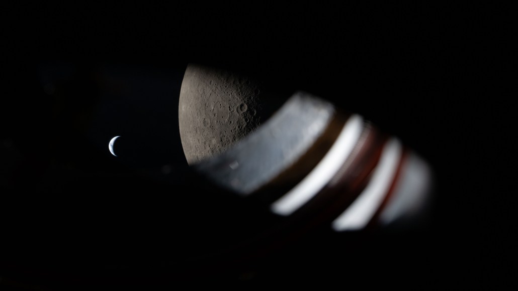 A view through the spacecraft window, wiht some of the window itself visible. The moon fills the right side of the frame and the smaller Earth is to the left.