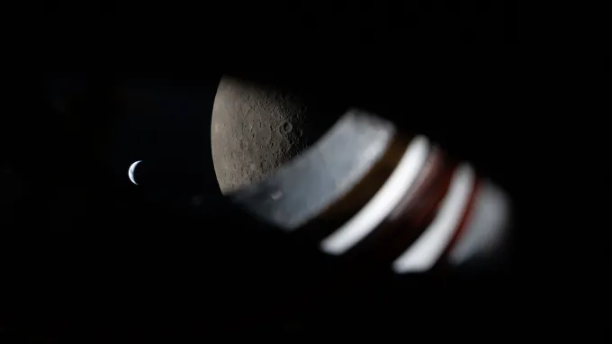 A view through the spacecraft window, wiht some of the window itself visible. The moon fills the right side of the frame and the smaller Earth is to the left.