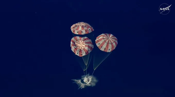 A spacecraft splashes down in the ocean beneath three large parachutes.