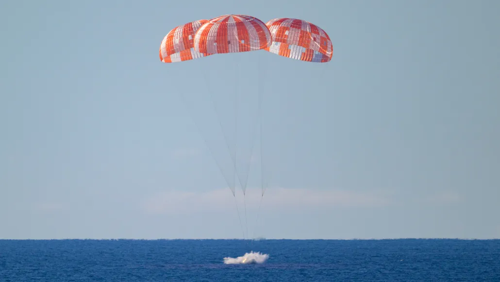 Three parachutes deploy above a spacecraft as it splashes into the ocean.