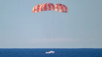 Three parachutes deploy above a spacecraft as it splashes into the ocean.