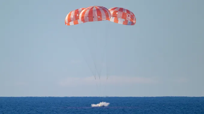 Three parachutes deploy above a spacecraft as it splashes into the ocean.
