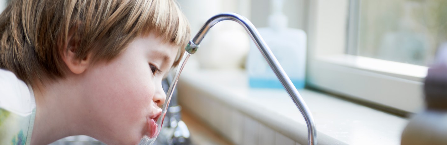 A little boy leans over a kitchen sink to drink water directly from the faucet.