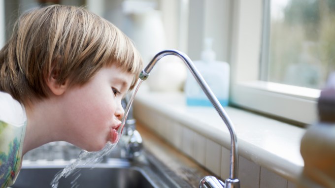 A little boy leans over a kitchen sink to drink water directly from the faucet.