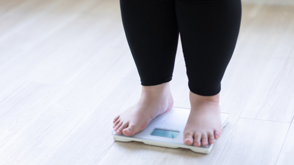 A woman wearing ankle-length black leggings stands barefoot on a scale. Her legs are visible below the knee only. Using GLP-1 drugs for obesity produces different results for people. Gene variants partly explain the drugs' efficacy and whether people will have side effects.