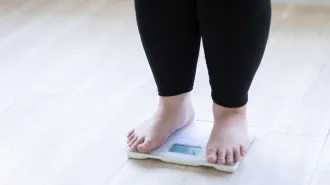 A woman wearing ankle-length black leggings stands barefoot on a scale. Her legs are visible below the knee only. Using GLP-1 drugs for obesity produces different results for people. Gene variants partly explain the drugs' efficacy and whether people will have side effects.
