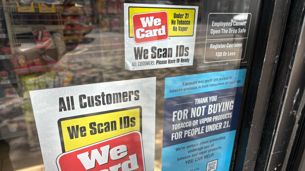 A convenience store window with posted signs explaining the legal age to buy tobacco products is 21.