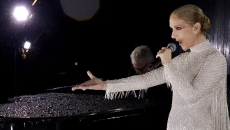 Celine Dion wears a beaded dress and stands holding a microphone with one hand outstretched while she performs at the Paris Olympics, A piano player and bright lights can be seen in the background.