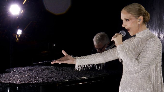 Celine Dion wears a beaded dress and stands holding a microphone with one hand outstretched while she performs at the Paris Olympics, A piano player and bright lights can be seen in the background.