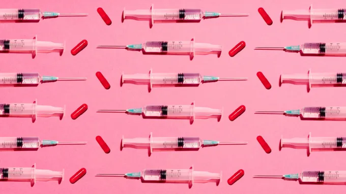 Partially filled syringes and red pill capsules are laid in a pattern on a pink background, in a way to illustrate how peptides may be administered.