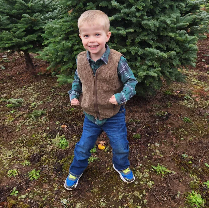 A smiling young boy with blond hair stands outside in front of a tree.