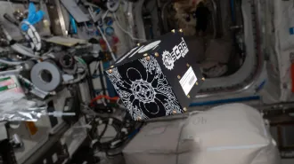 A black and white cube with the name "OSCAR QUBE" on one side floats in the International Space Station.