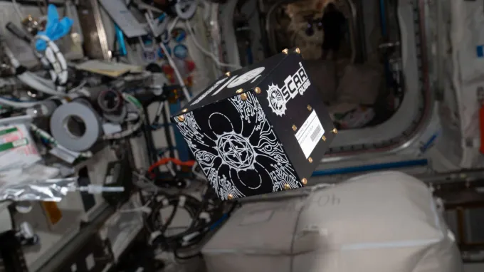 A black and white cube with the name "OSCAR QUBE" on one side floats in the International Space Station.