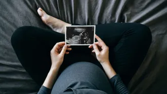 A pregnant person sits cross legged on a bed holding an ultrasound image of the fetus.