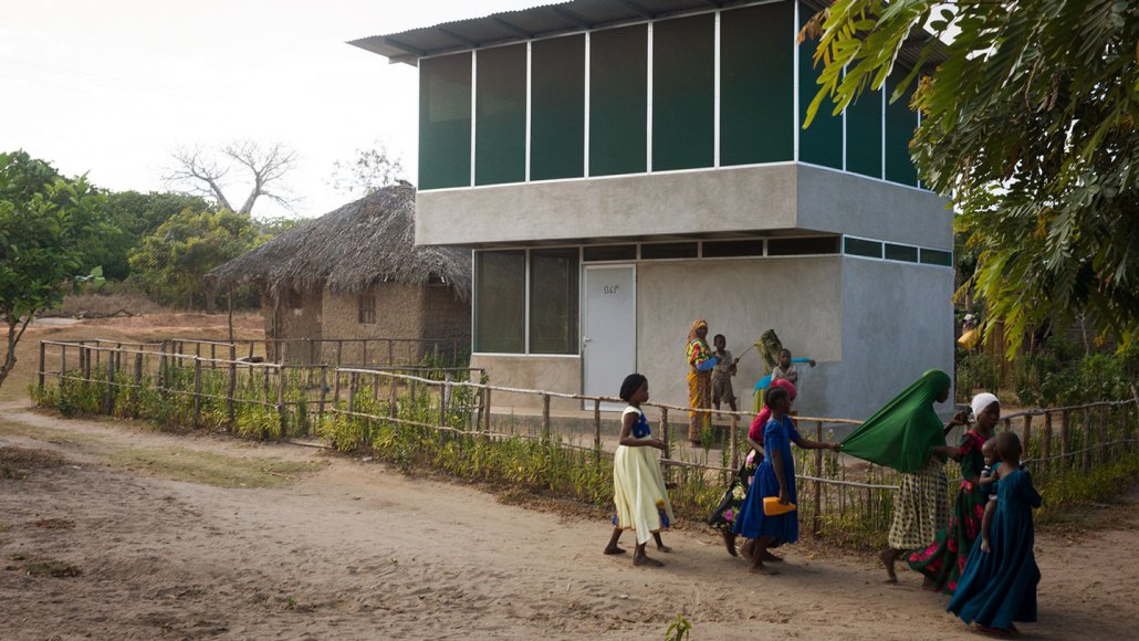 Children walk along a dirt road in Tanzania past a two-story house with screened openings. A more traditional one story house with a thatched roof can be seen next door.