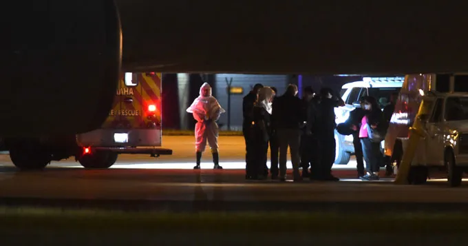 A cluster of people can be seen silhouetted under the belly of a plane while waiting to board a van. A person in full white protective gear stands to the left looking on. These are American passengers from the MV Hondius cruise ship medically evacuated to Omaha, Nebraska, for monitoring or treatment for hantavirus infection.
