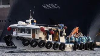 A small white boat pulls away from the black hull of a ship on which the name Hondius is visible in white letters. Eight people in blue medical garb with white showercaps on their heads and wearing N95 masks sit in the back of the boat. A person in a full body white suit including a hood and N95 mask is standing at the front left bow while another person in full white protective gear is in the cabin. These are American citizens being evacuated from a cruise ship after an Andes hantavirus outbreak onboard.