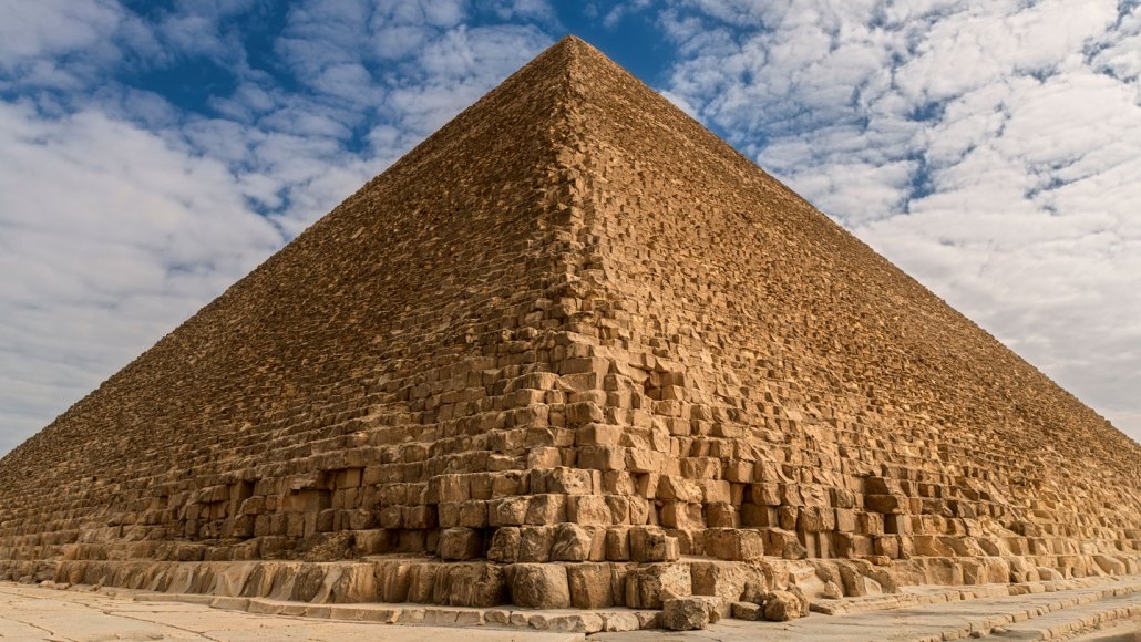 Egypt's Great Pyramid, seen looking up from the vantage point at one of its corners. The sand-colored stones rise up against a cloudy blue sky background.