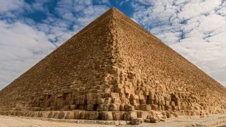Egypt's Great Pyramid, seen looking up from the vantage point at one of its corners. The sand-colored stones rise up against a cloudy blue sky background.