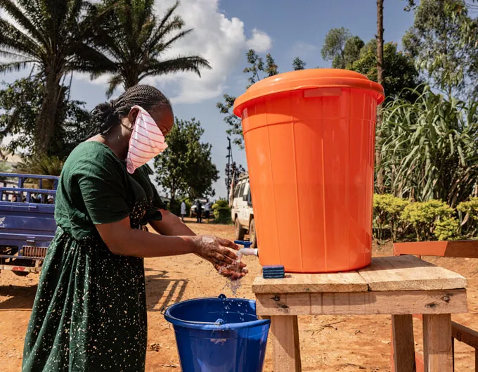 A woman wearing a scarf over her face washes her hands at an outdoor stand holding water in a big orange bucket, with a blue bucket to catch the used water.