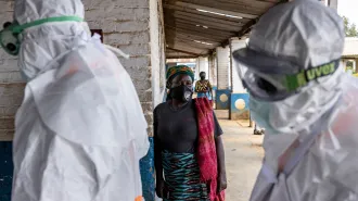In the foreground, two people completely covered in white protective gear and goggles walk by two women who are wearing face masks.