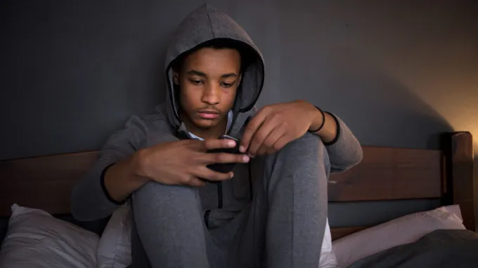 A teenager in a gray sweatshirt and sweatpants with a sad expression sits on a bed looking at a smartphone screen