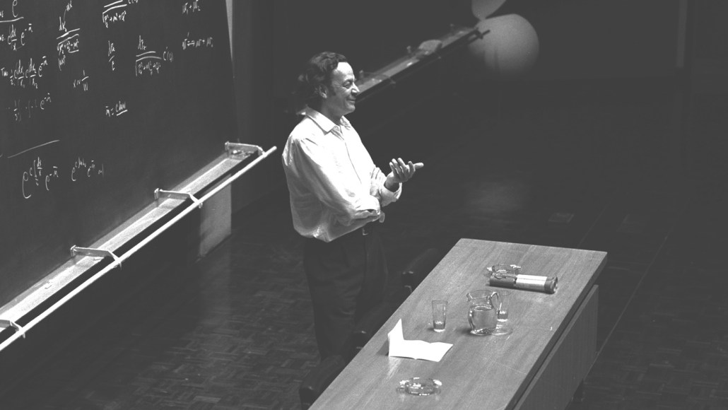 Richard Feynman stands in front of a blackboard in a lecture hall.