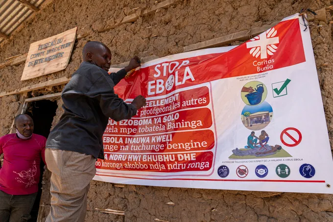 A person hands a large poster that reads "STOP EBOLA" on the side of a mud-constructed building