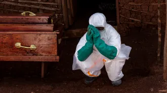A health care worker wearing a full-body, white biohazard suit with teal gloves that come up to the elbow crouches with their hands with their hands held in front of their face, almost as if in prayer. A coffin is to the worker's right.