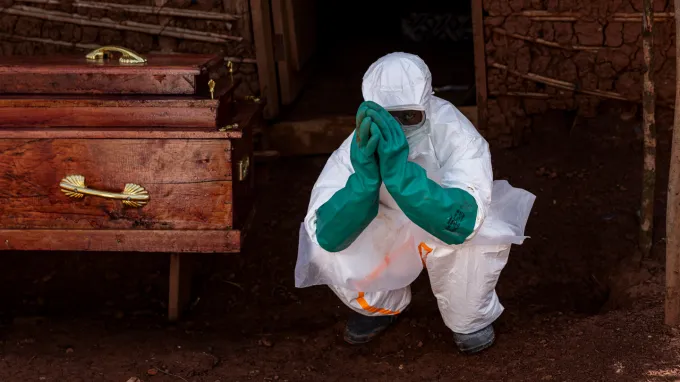 A health care worker wearing a full-body, white biohazard suit with teal gloves that come up to the elbow crouches with their hands with their hands held in front of their face, almost as if in prayer. A coffin is to the worker's right.