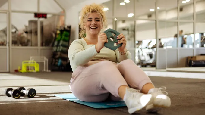 A smiling woman seated in a gym holding a weight.
