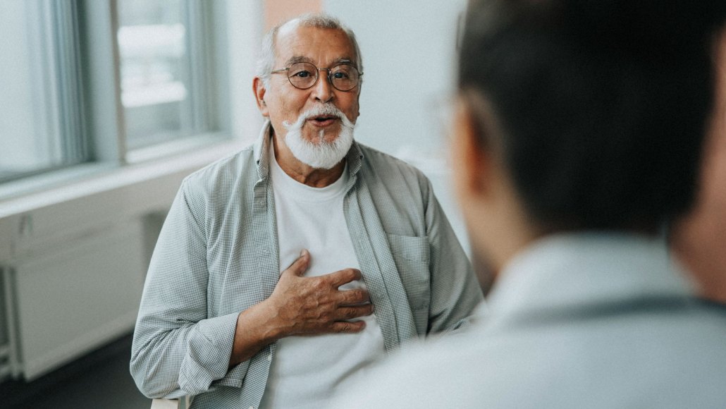 An older man with a white mustache and beard, wearing glasses and with his hand over his heart talks with a medical provider in an office.