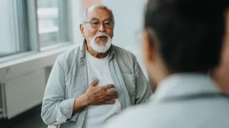 An older man with a white mustache and beard, wearing glasses and with his hand over his heart talks with a medical provider in an office.