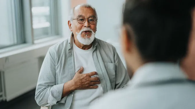 An older man with a white mustache and beard, wearing glasses and with his hand over his heart talks with a medical provider in an office.