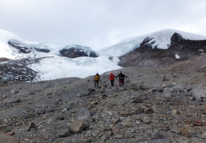 Three people in cold-weather gear traverse a boulder-strewn slope, with snow-covered glaciers visible behind them.