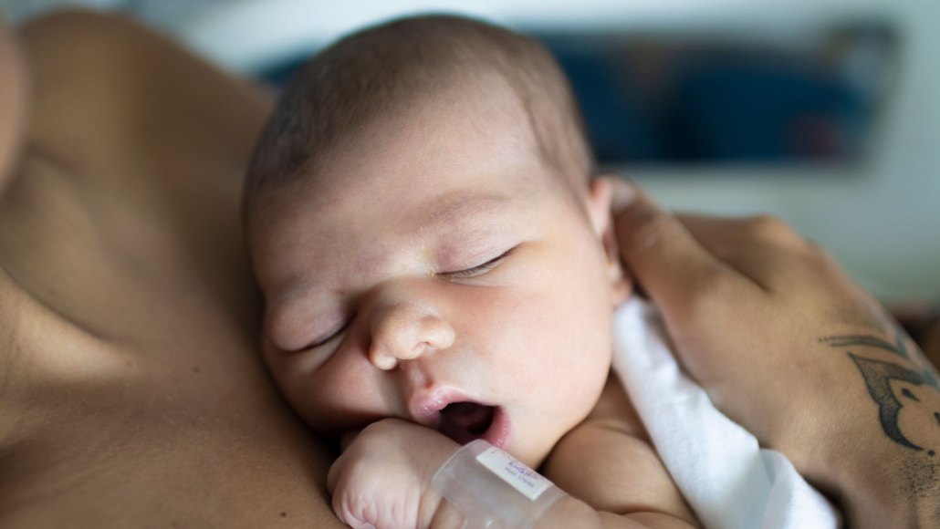 a newborn baby, with eyes closed and mouth open, rests on their parent's chest