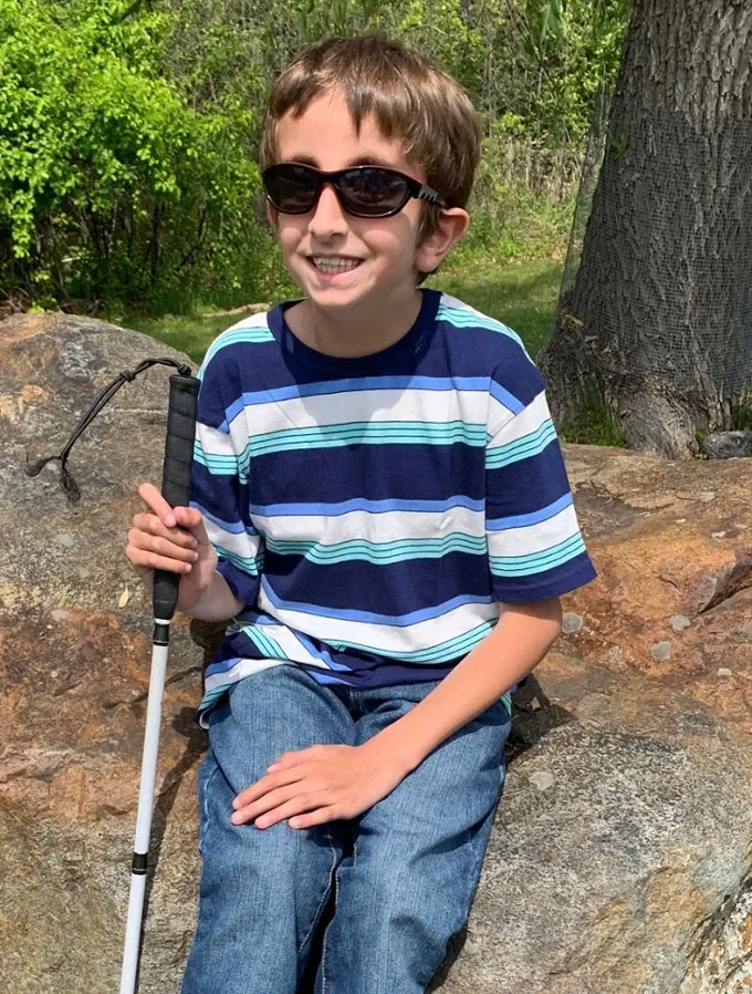 A boy in a striped shirt wearing black sunglasses and holding a white walking cane.